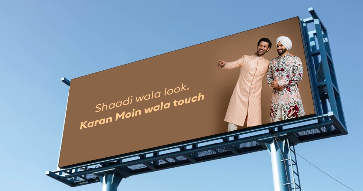 A large roadside billboard against a blue sky displays a wedding fashion advertisement. Two men wearing traditional Indian wedding outfits stand on the right side of the hoarding, while the headline “Shaadi wala look. Karan Main wala touch” appears prominently on a brown background. The ad demonstrates how concise copy and strong visuals work together to communicate a brand message instantly.